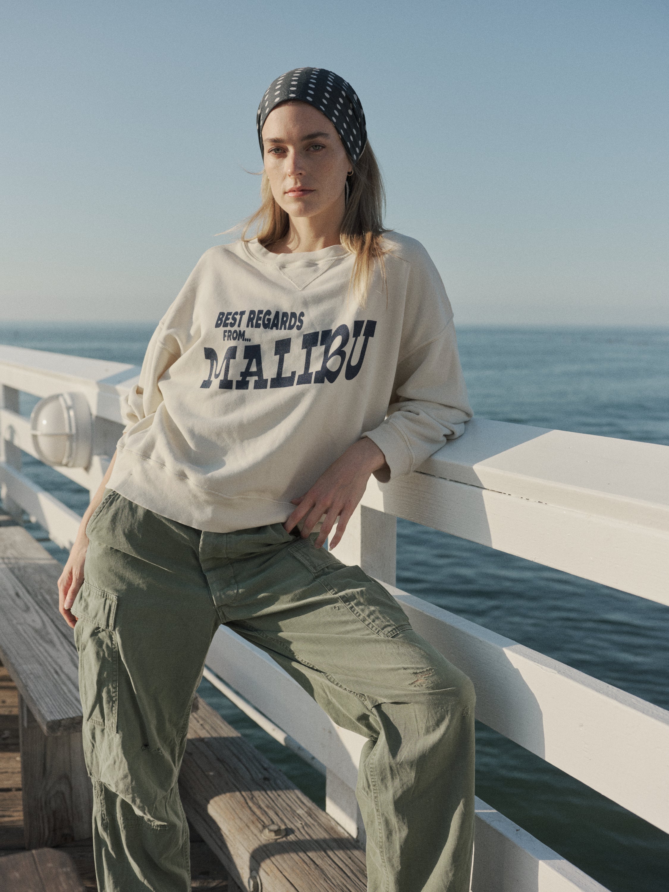 Person wearing a 'Best Regards from Malibu' sweatshirt sitting on a railing with ocean view