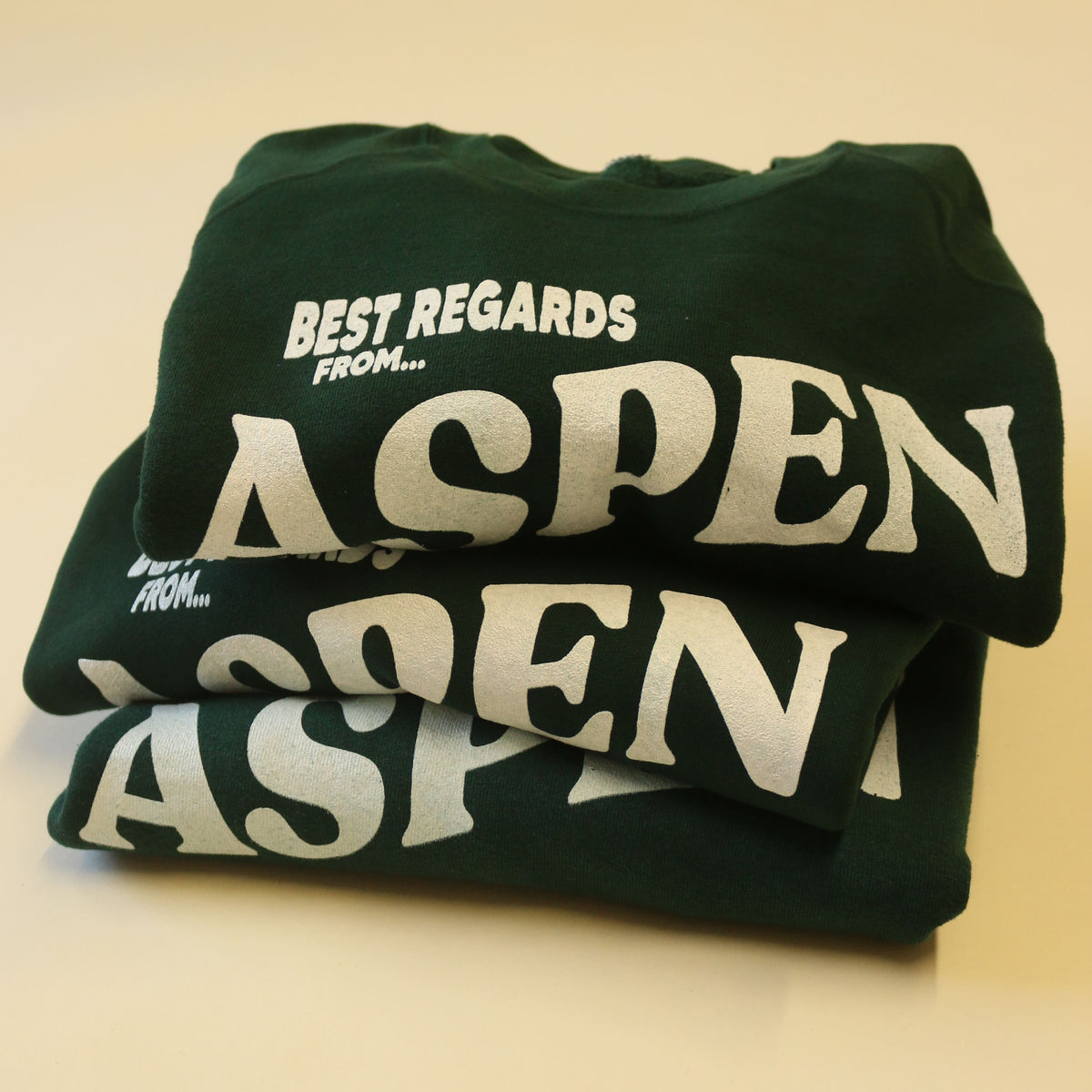 A folded dark green t-shirt with large white 'ASPEN' lettering and 'BEST REGARDS FROM.